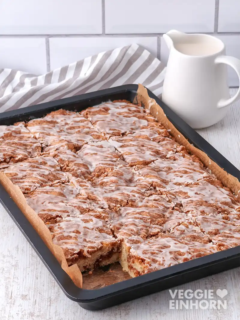 Zimtkuchen mit Zuckerguss auf einem Backblech, im Hintergrund ein Küchentuch und ein Krug mit veganer Milch.