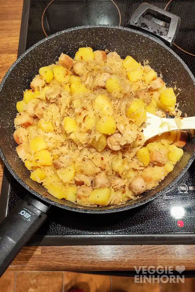 Eine große Pfanne auf dem Herd, gefüllt mit Sauerkraut, Kartoffelwürfeln und gebratenen veganen Bratwurststücken.