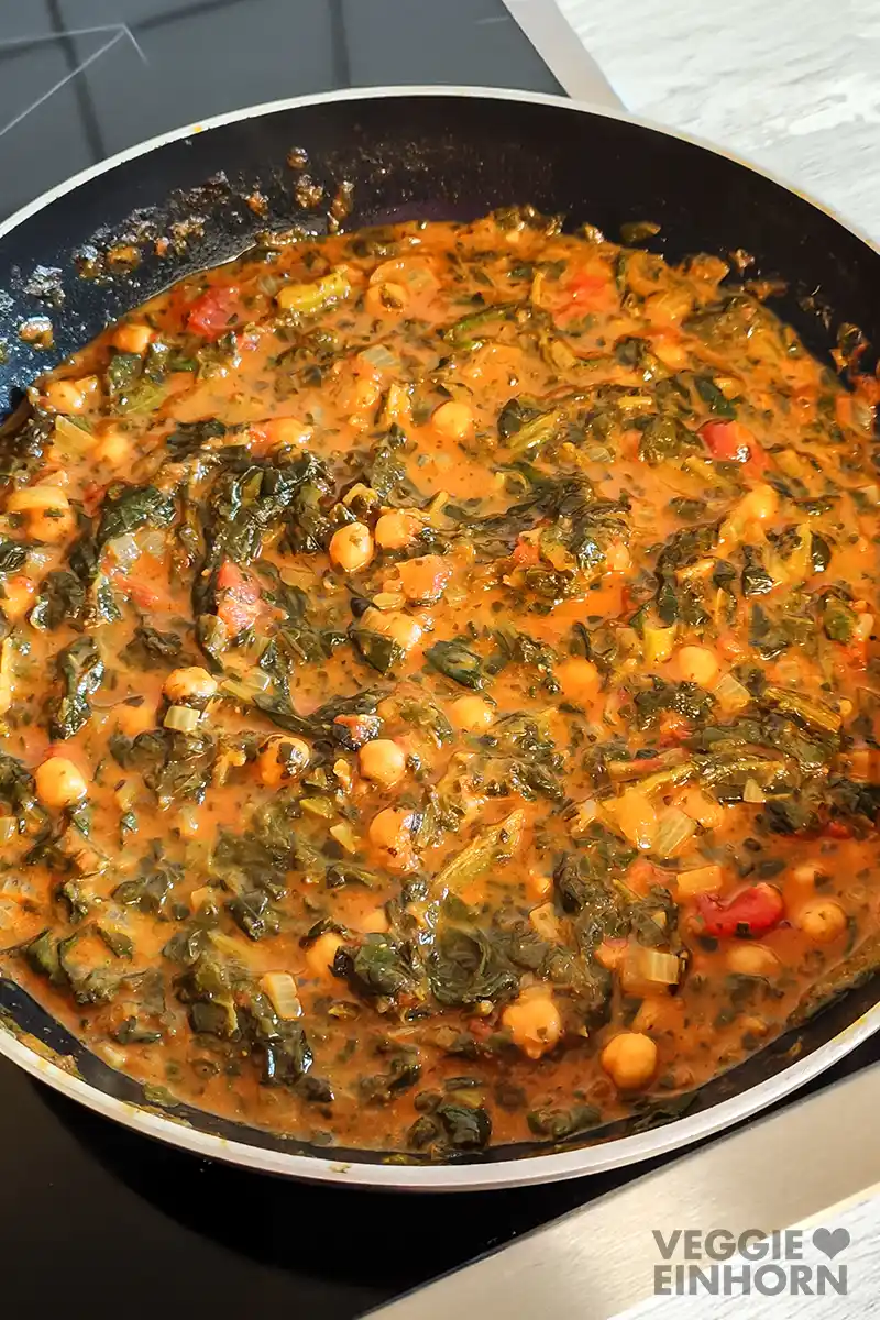 Curry aus Spinat, Kichererbsen, Tomaten und Kokosmilch in einer schwarzen Pfanne auf dem Kochfeld.