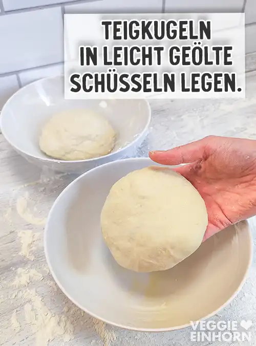Eine Hand legt eine runde Teigkugel in eine weiße Schüssel, im Hintergrund steht eine weitere Schüssel mit Teigkugel. Auf dem Bild steht der Text Teigkugeln in leicht geölte Schüsseln legen.