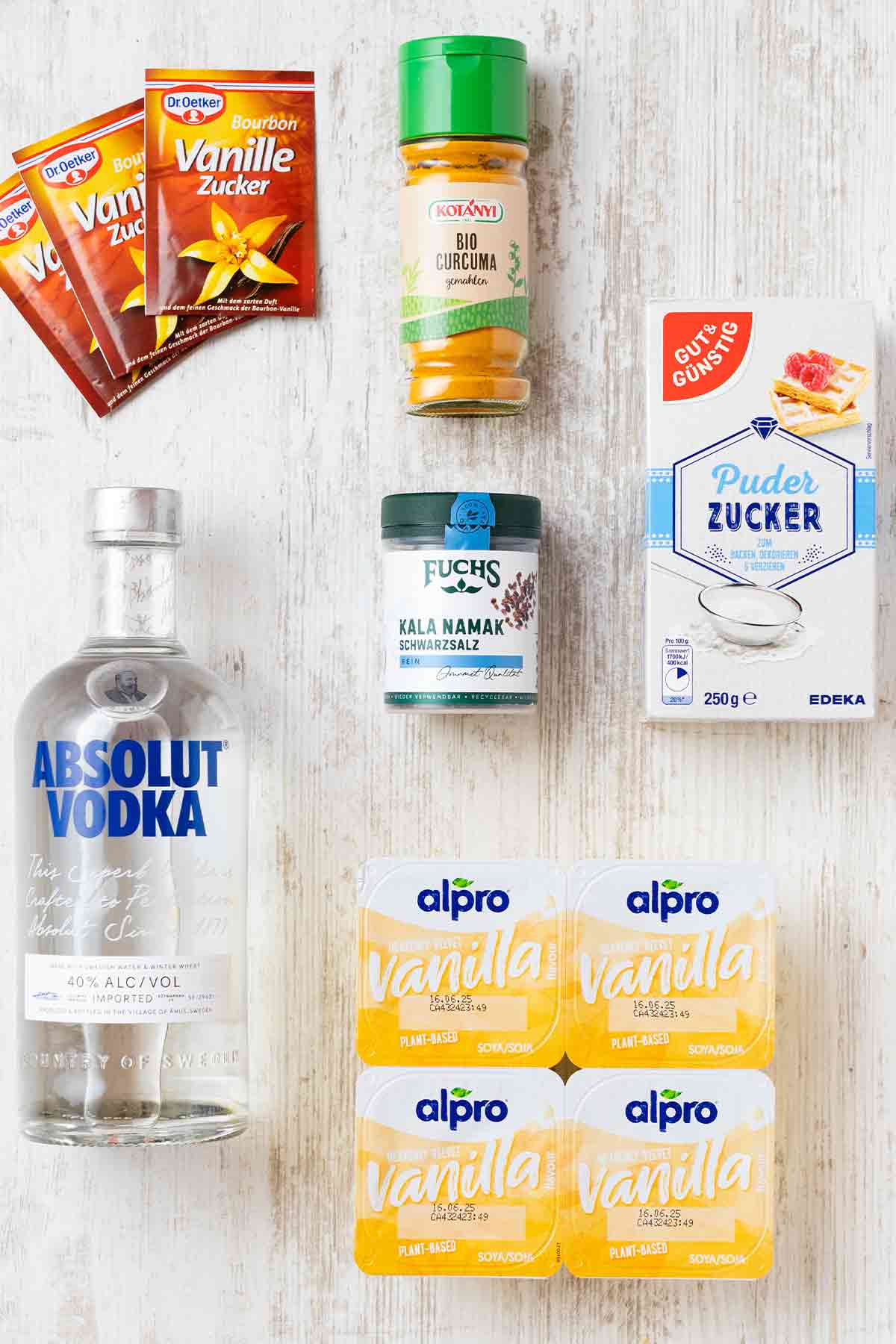 Zutaten für veganen Eierlikör auf einem weißen Holztisch: Vanillepudding, Wodka, Puderzucker, Vanillezucker, Kurkuma, Kala Namak.