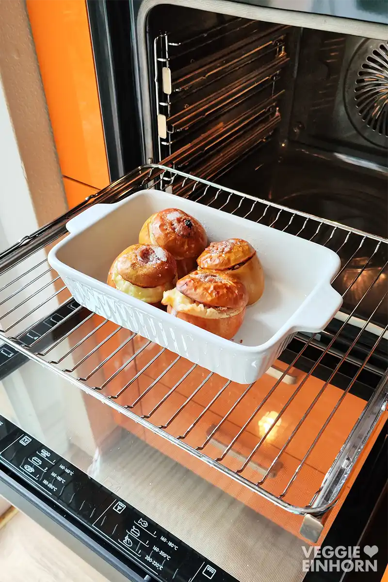 Vier gefüllte Bratäpfel in einer weißen Auflaufform stehen im geöffneten Backofen und sind goldbraun gebacken.