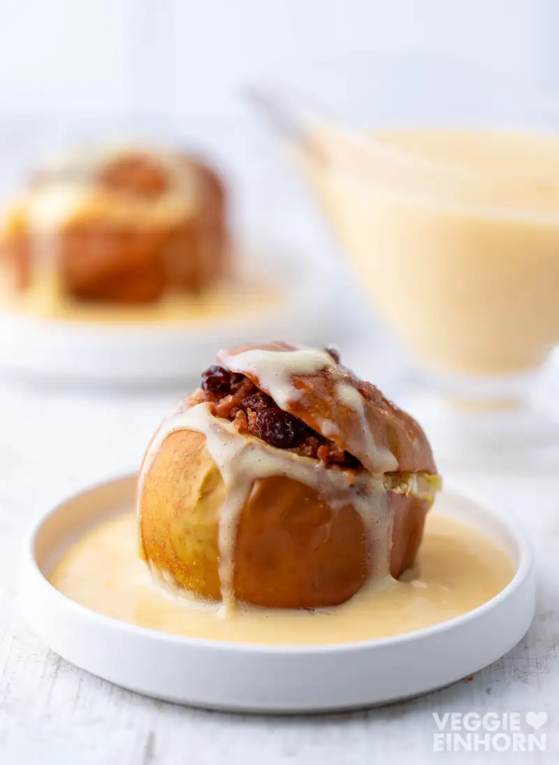 Ein Bratapfel mit veganer Vanillesoße und Rosinenfüllung auf einem weißen Teller, im Hintergrund unscharf eine Sauciere.