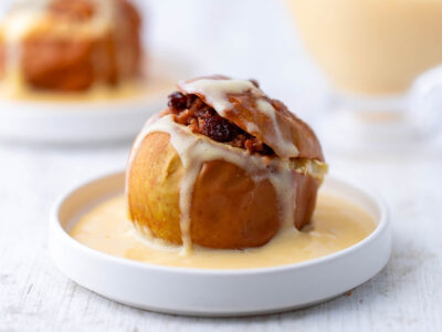 Ein veganer Bratapfel mit Rosinen- und Spekulatiusfüllung auf einem weißen Teller mit veganer Vanillesoße.