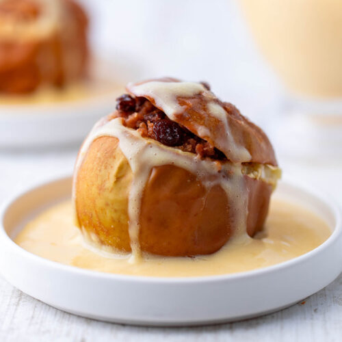 Ein veganer Bratapfel mit Rosinen- und Spekulatiusfüllung auf einem weißen Teller mit veganer Vanillesoße.