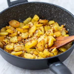 Vegane Bratkartoffeln mit Räuchertofu und Zwiebeln in einer schwarzen Pfanne mit einem Holzlöffel.