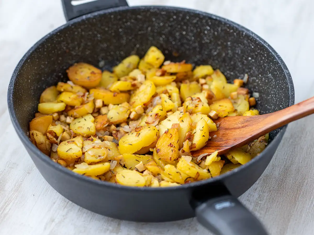 Vegane Bratkartoffeln mit Räuchertofu und Zwiebeln in einer schwarzen Pfanne mit einem Holzlöffel.
