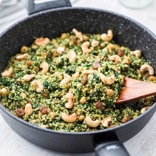 Eine schwarze Pfanne gefüllt mit einer veganen Couscous-Pfanne, die mit Kichererbsen, Spinat, Cashewkernen und Rosinen zubereitet ist. Ein Holzlöffel rührt das bunte Gericht um.