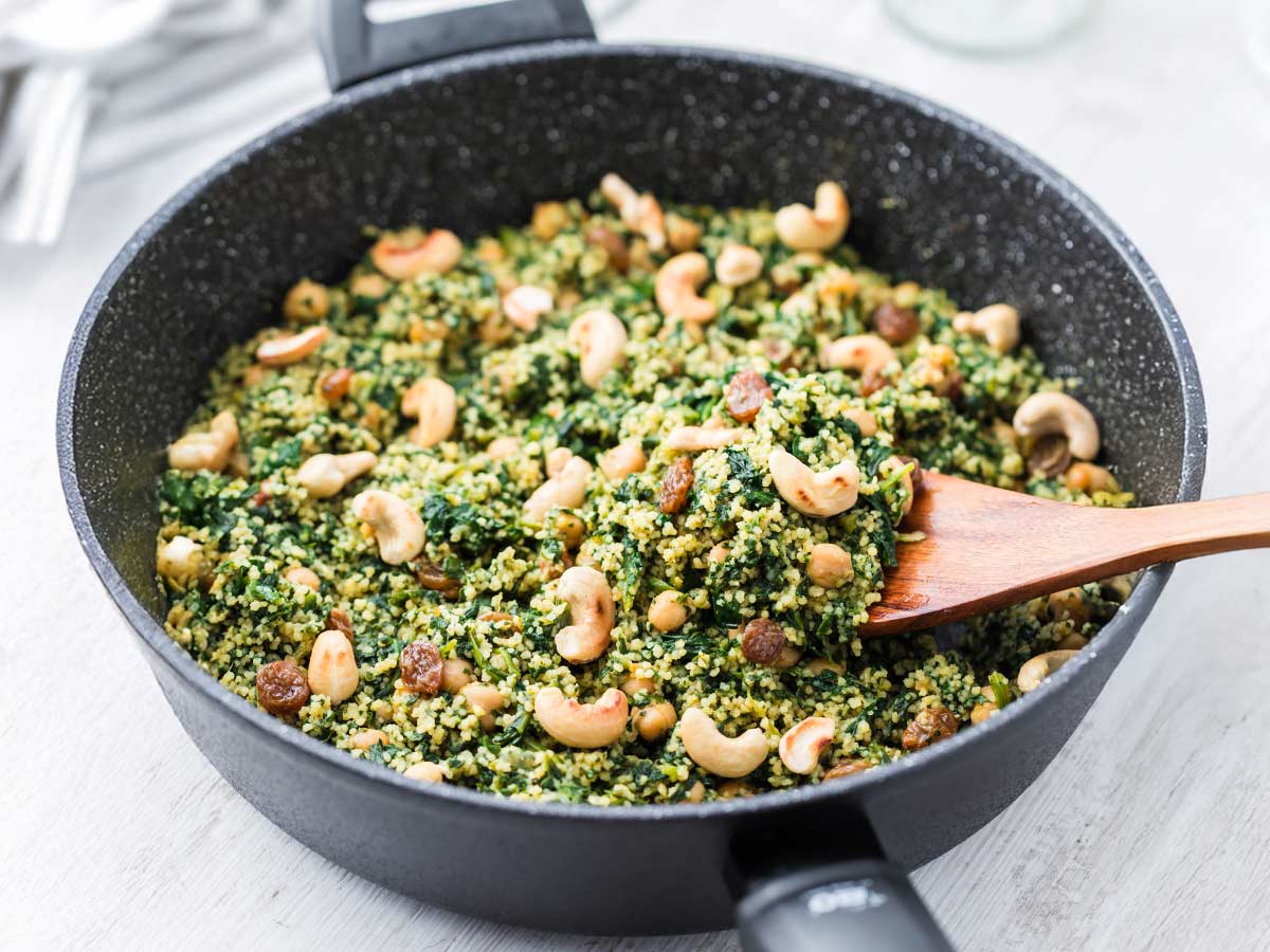 Eine schwarze Pfanne gefüllt mit einer veganen Couscous-Pfanne, die mit Kichererbsen, Spinat, Cashewkernen und Rosinen zubereitet ist. Ein Holzlöffel rührt das bunte Gericht um.