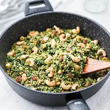 Eine schwarze Pfanne gefüllt mit einer veganen Couscous-Pfanne, die mit Kichererbsen, Spinat, Cashewkernen und Rosinen zubereitet ist. Ein Holzlöffel rührt das bunte Gericht um.