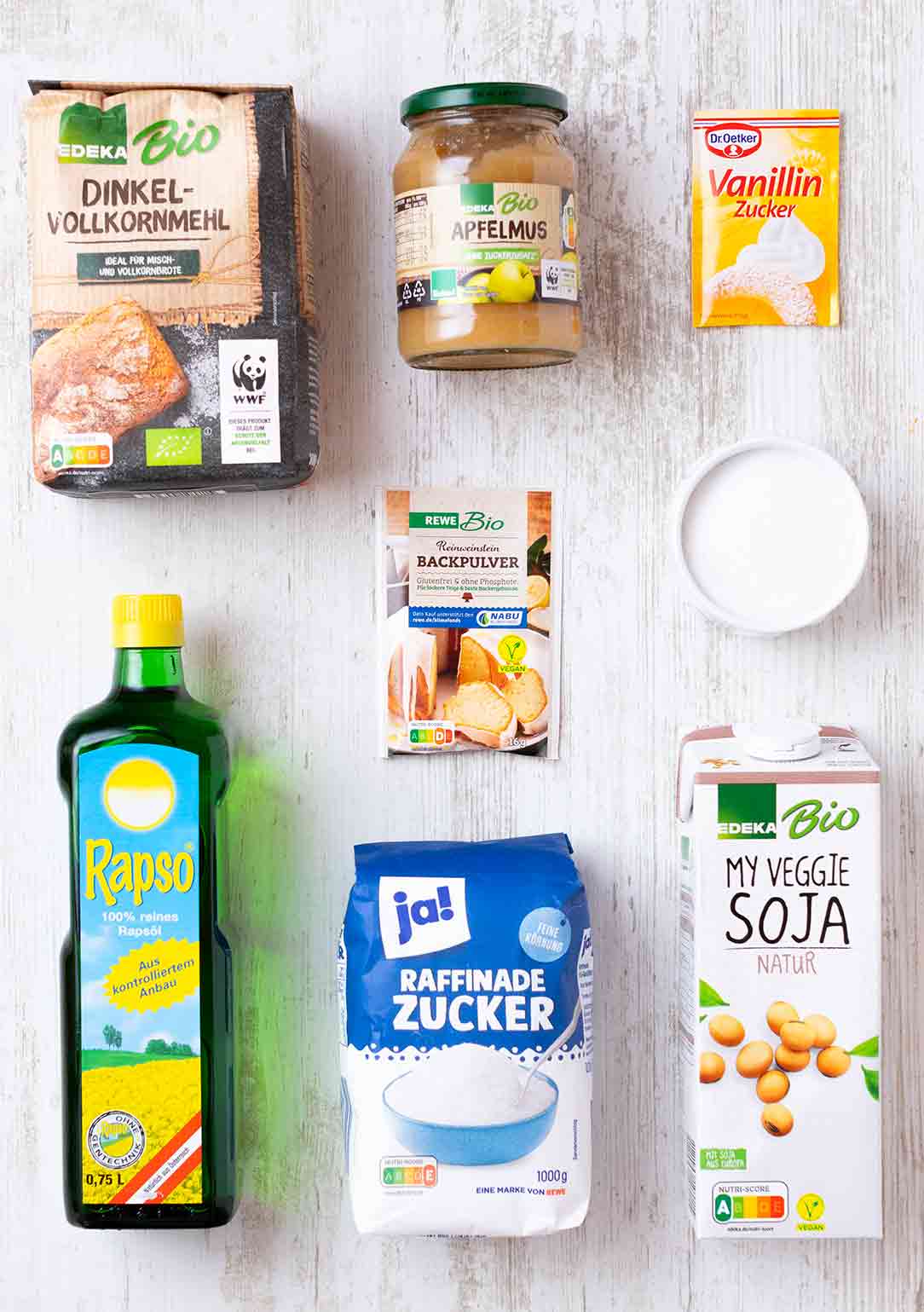 Zutaten für vegane Dinkelwaffeln auf einem weißen Holztisch: Mehl, Apfelmus, Sojamilch, Zucker, Öl, Vanillezucker, Backpulver.