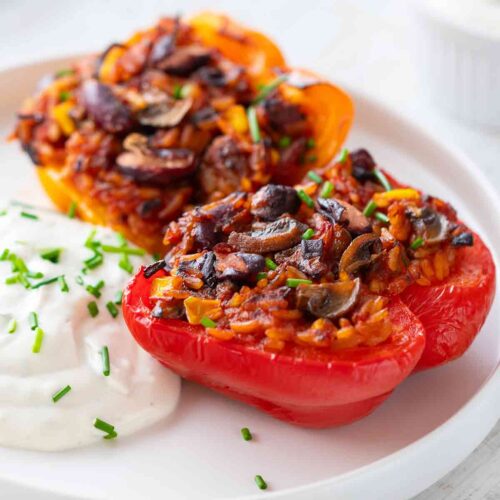Zwei Hälften veganer gefüllter Paprika mit einer würzigen Reis-Gemüse-Mischung, serviert auf einem weißen Teller mit einem Klecks veganem Kräuterquark, bestreut mit frischem Schnittlauch.