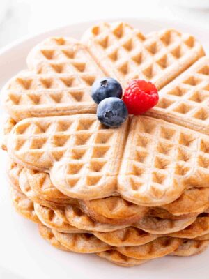 Nahaufnahme von herzförmigen veganen Vollkornwaffeln ohne Zucker, mit frischen Beeren dekoriert, auf einem weißen Teller serviert.