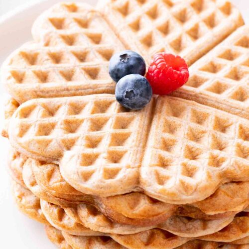Nahaufnahme von herzförmigen veganen Vollkornwaffeln ohne Zucker, mit frischen Beeren dekoriert, auf einem weißen Teller serviert.
