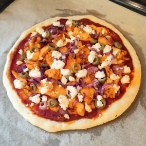 Vegane Gyrospizza auf einem Backblech mit Tomatensauce, veganen Gyrosstücken, roten Zwiebeln, Oliven und zerbröckeltem veganem Feta.