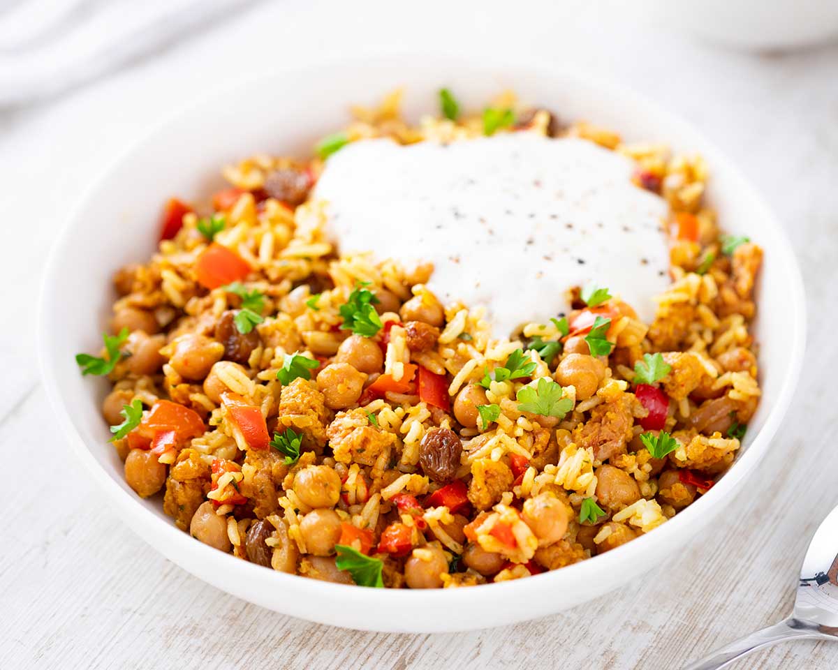Weißer Teller mit veganer Hackfleisch-Reis-Pfanne aus Kichererbsen, Paprika, veganem Hack, Rosinen, Reis und Joghurt auf einem weißen Tisch.