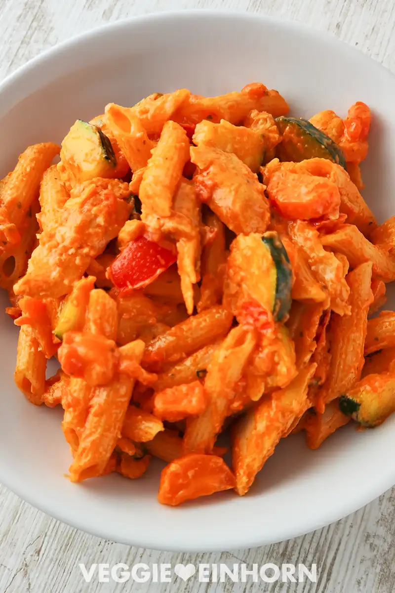 Weißer Teller mit cremiger veganer Hähnchenpfanne mit Nudeln, Zucchini, Möhren und Tomaten.