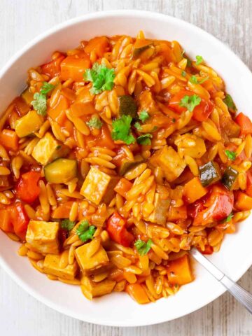 Nahaufnahme einer veganen Kritharaki-Pfanne mit Räuchertofu, Paprika, Zucchini und Möhren in einer aromatischen Tomatensoße, serviert in einem weißen Teller und garniert mit frischer Petersilie.