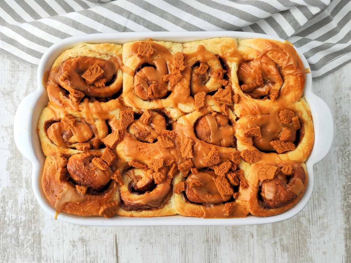 Vegane Lotus Zimtschnecken in einer weißen Form, goldbraun gebacken und mit karamellfarbener Biscoff Glasur überzogen. Obenauf liegen knusprige Lotuskeks Stückchen.