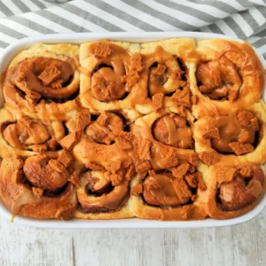 Goldbraune vegane Lotus Zimtschnecken in einer weißen Auflaufform, mit cremiger Biscoff Glasur und zerbröselten Lotus Keksen bestreut.