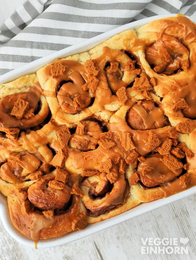 Vegane Lotus Zimtschnecken mit cremiger Lotus Biscoff Glasur in einer weißen Auflaufform. Obenauf liegen knusprige Lotus Keksstücke, im Hintergrund ein gestreiftes Küchentuch.