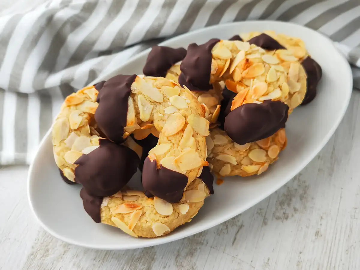 Vegane Mandelhörnchen auf einem weißen Teller, mit Mandelblättchen bedeckt und in dunkle Schokolade getaucht.
