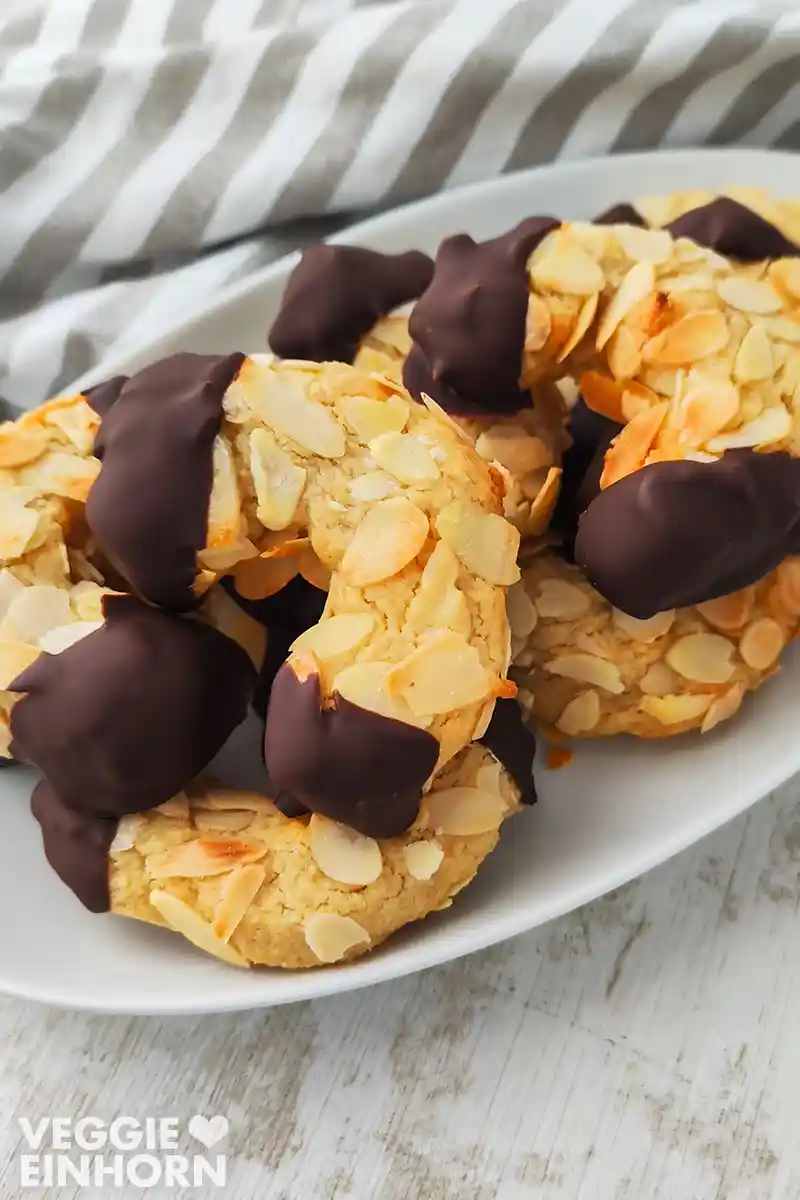 Mehrere vegane Mandelhörnchen auf einem weißen Teller, goldbraun gebacken und mit dunkler Schokolade verziert.