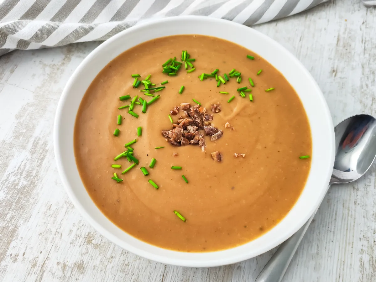 Cremige vegane Kartoffel-Maronensuppe in einem weißen Teller, garniert mit frischem Schnittlauch und kleinen Stücken Maronen auf hellem Holztisch.
