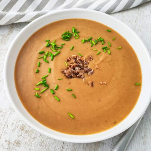 Vegane Maronen-Kartoffelsuppe in einem weißen Teller mit grünen Schnittlauchringen und gehackten Maronen, daneben ein Löffel auf hellem Holzuntergrund.