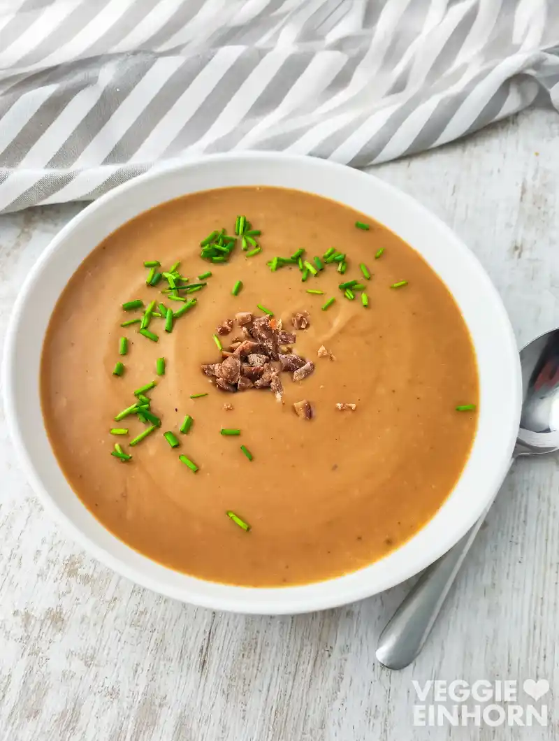 Teller mit veganer Kartoffel-Maronensuppe auf einem hellen Holztisch, daneben liegen eine gestreifte Stoffserviette und ein Löffel.