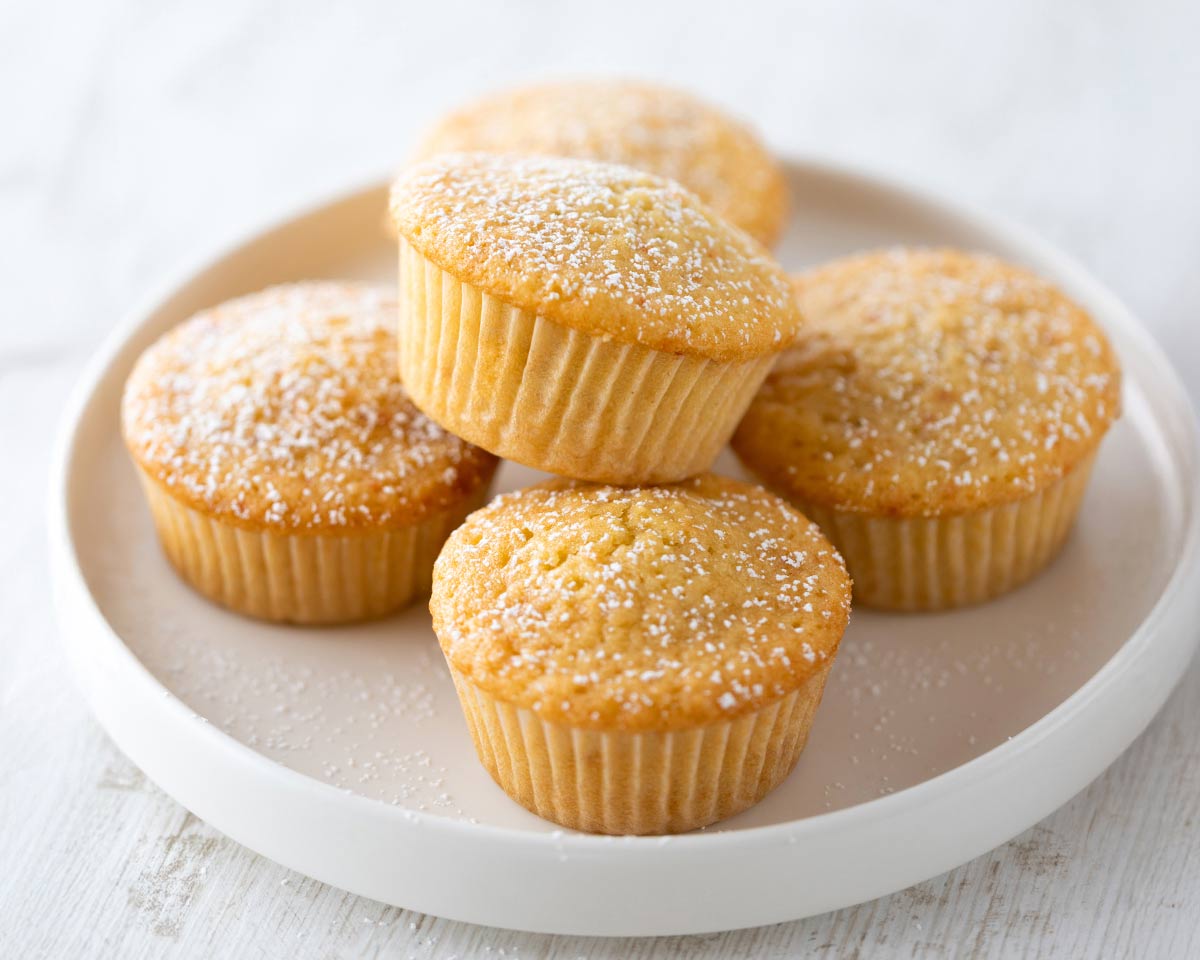 Fluffige vegane Muffins in Papierförmchen, mit Puderzucker bestäubt, auf einem weißen Teller.