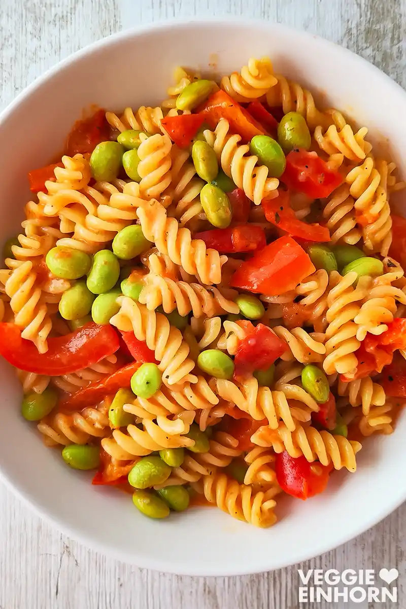 Nahaufnahme einer veganen Nudelpfanne mit Edamame, Paprika und Tomaten, serviert auf einem weißen Teller.