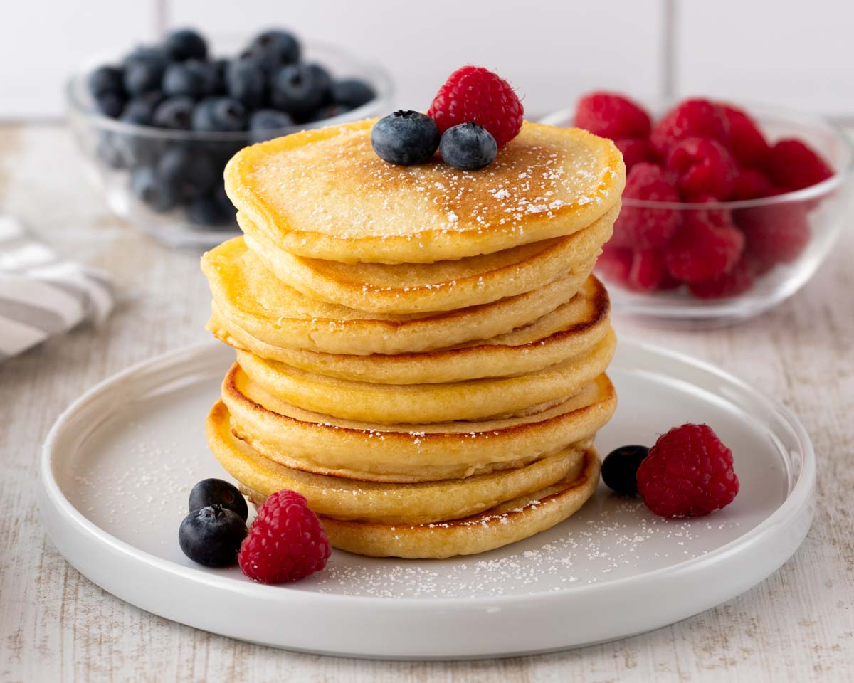 Ein Stapel goldbrauner veganer Pancakes, garniert mit frischen Himbeeren und Blaubeeren, serviert auf einem weißen Teller. Im Hintergrund befinden sich Schalen mit zusätzlichen Beeren.