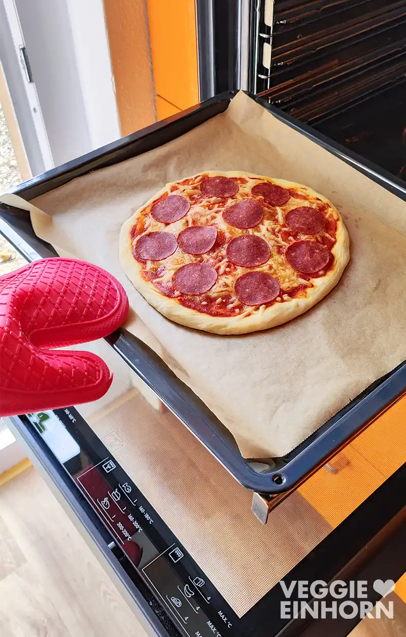 Vegane Pizza Salami frisch aus dem Ofen auf einem Backblech mit Backpapier, gehalten von einer Hand mit rotem Ofenhandschuh.