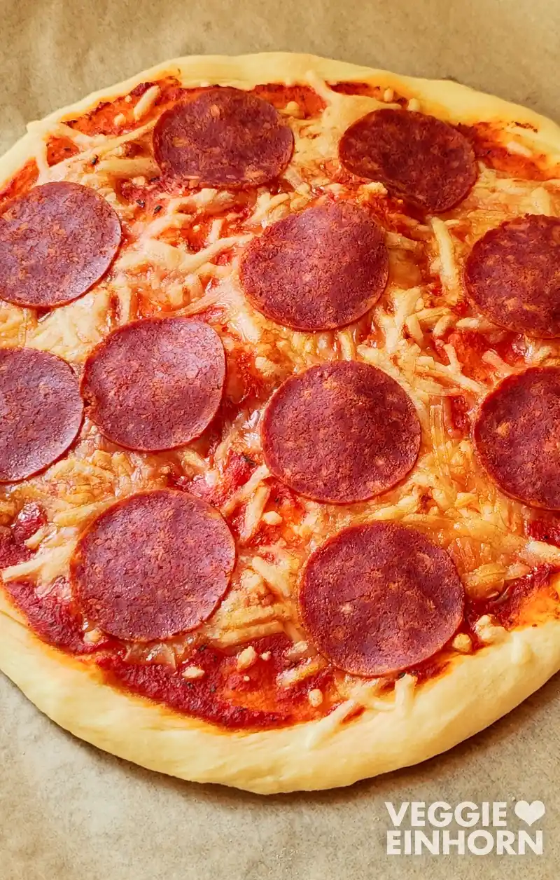 Nahaufnahme einer runden veganen Pizza mit Tomatensauce, geschmolzenem Käse und mehreren Scheiben veganer Salami auf goldbraun gebackenem Teig.
