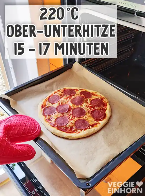 Vegane Pizza Salami auf einem Backblech mit Backpapier wird mit einem roten Ofenhandschuhaus dem Ofen gezogen, darüber Textaufschrift 220 °C Ober-Unterhitze 15–17 Minuten.