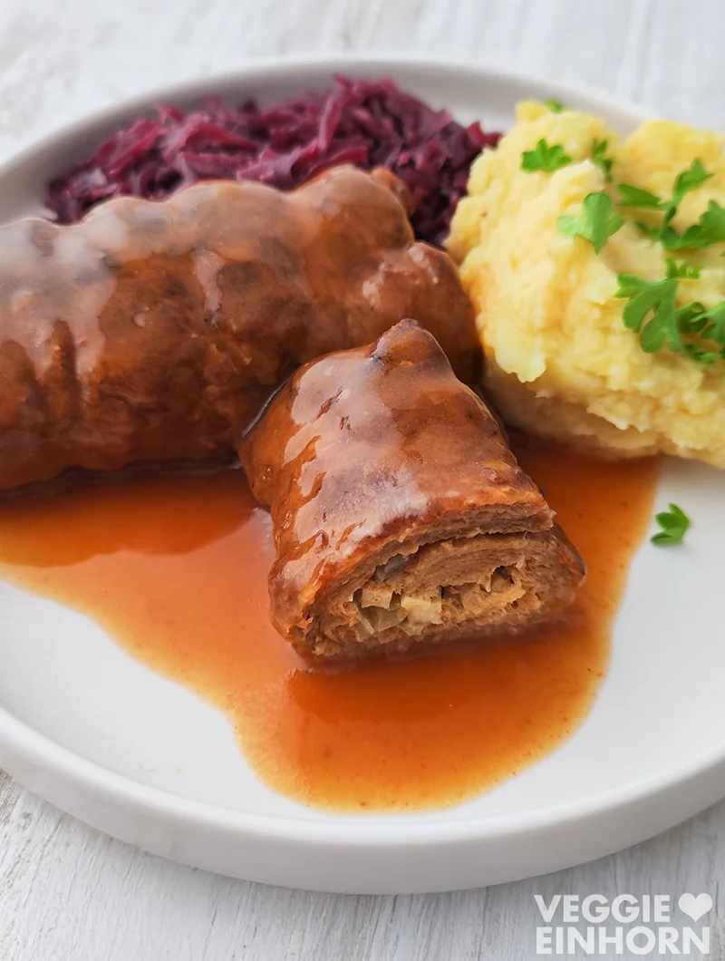 Vegane Roulade aus Soja Big Steaks mit veganer Bratensoße, Rotkohl und Kartoffelpüree auf einem weißen Teller.