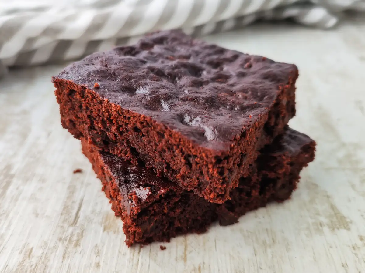 Zwei vegane Schoko Brownies liegen übereinander auf einem hellen Holztisch mit einem gestreiften Tuch im Hintergrund.