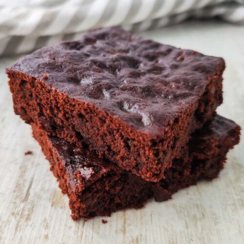 Zwei übereinander gestapelte vegane Schokobrownies auf heller Holzoberfläche mit gestreiftem Tuch im Hintergrund.