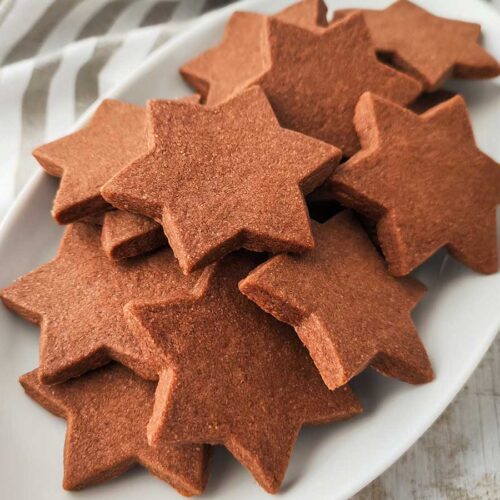 Mehrere sternförmige vegane Schokoplätzchen liegen auf einem weißen Teller vor einem gestreiften Tuch.