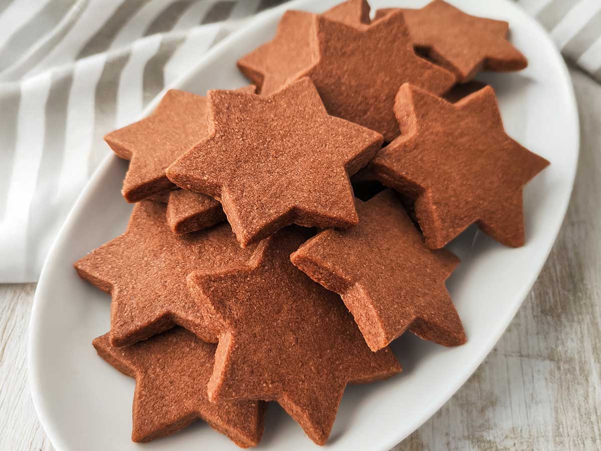 Mehrere sternförmige vegane Schokoplätzchen liegen auf einem weißen Teller vor einem gestreiften Tuch.