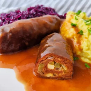 Vegane Seitan Rouladen liegen in brauner Soße auf einem Teller, eine Roulade ist angeschnitten und zeigt die Füllung aus Gewürzgurken, Räuchertofu und Zwiebeln, dazu Kartoffelpüree und Rotkohl im Hintergrund.