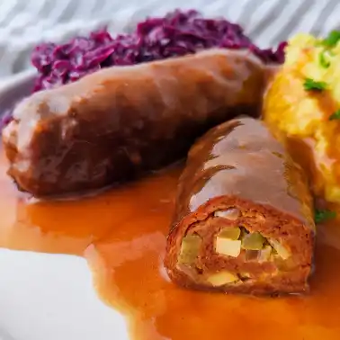 Zwei vegane Seitanrouladen in brauner Soße auf einem weißen Teller mit Kartoffelpüree und Rotkohl, eine Roulade ist angeschnitten und zeigt die Füllung.