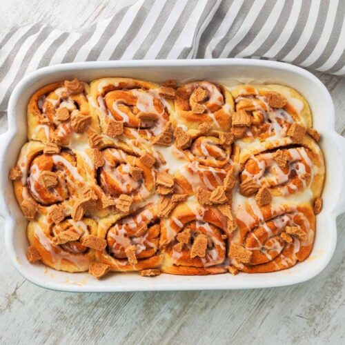 Weiße Auflaufform mit veganen Spekulatius Zimtschnecken auf hellem Holztisch mit einem gestreiftem Küchentuch.