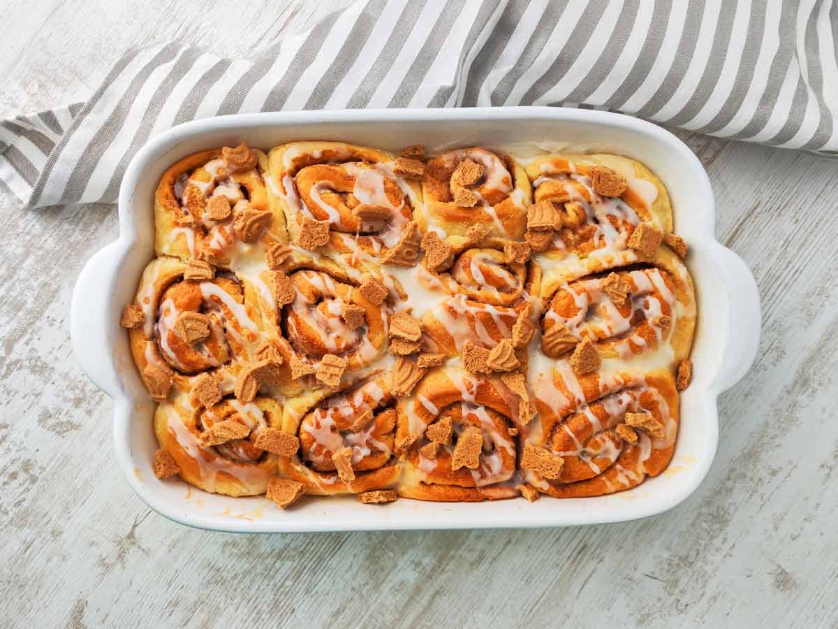 Weiße Auflaufform mit veganen Spekulatius Zimtschnecken auf hellem Holztisch mit einem gestreiftem Küchentuch.