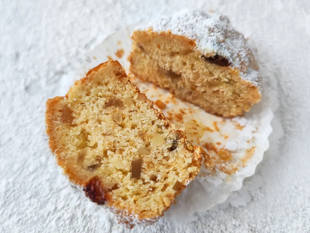 Halbierter veganer Stollen Muffin im Papierförmchen auf heller, mit Puderzucker bestäubter Fläche.