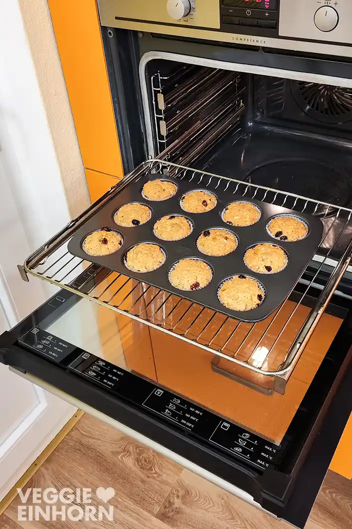 Muffinform mit 12 veganen Stollenmuffins auf dem Backofenrost vor dem geöffneten Backofen.