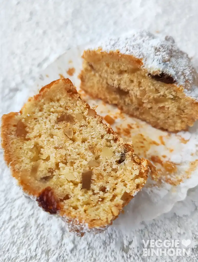 Nahaufnahme eines aufgeschnittenen veganen Stollen Muffins im Papierförmchen auf mit Puderzucker bestäubter Oberfläche.
