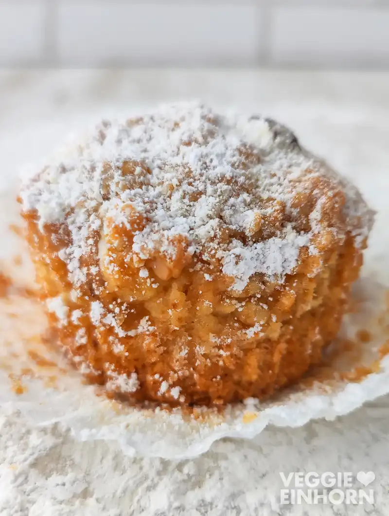 Ein veganer Stollenmuffin im Papierförmchen, bestäubt mit Puderzucker auf heller Unterlage.