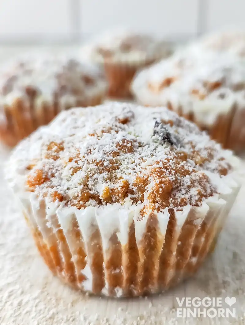Veganer Stollenmuffin mit Puderzucker im Papierförmchen auf weißer, leicht bestäubter Unterlage. Im Hintergrund stehen weitere Muffins.